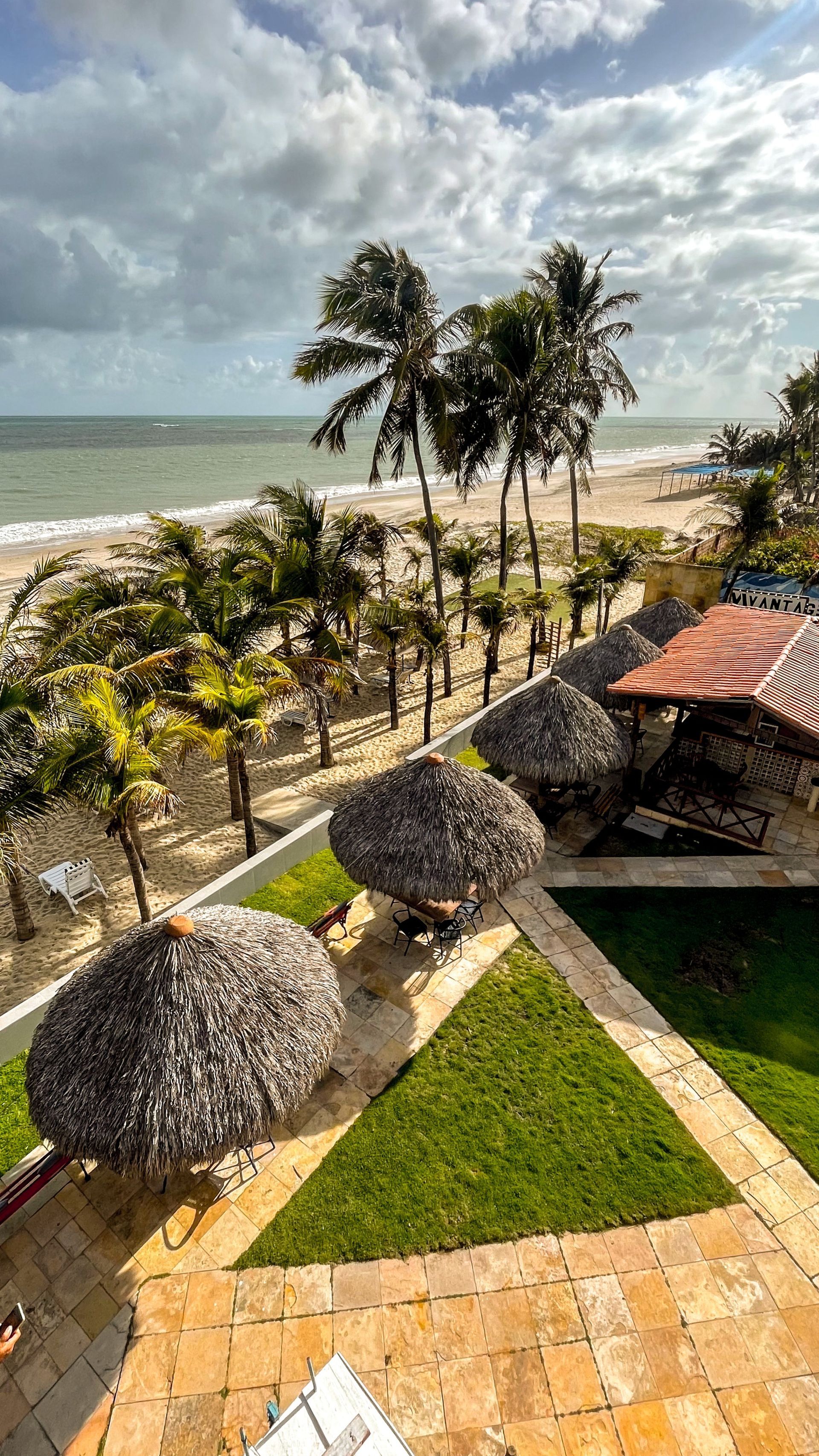 Jardim de resort costeiro com palmeiras, arbustos podados, caminho de pedra e oceano sob um céu nublado.