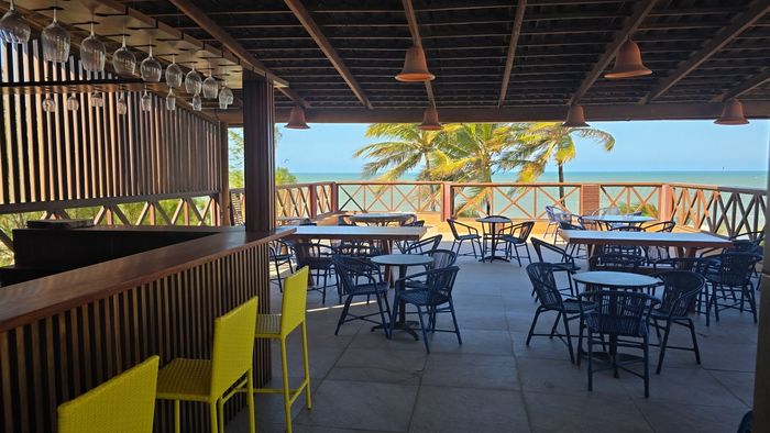 Café ao ar livre à beira-mar com mesas, cadeiras e vista para o oceano além do parapeito.