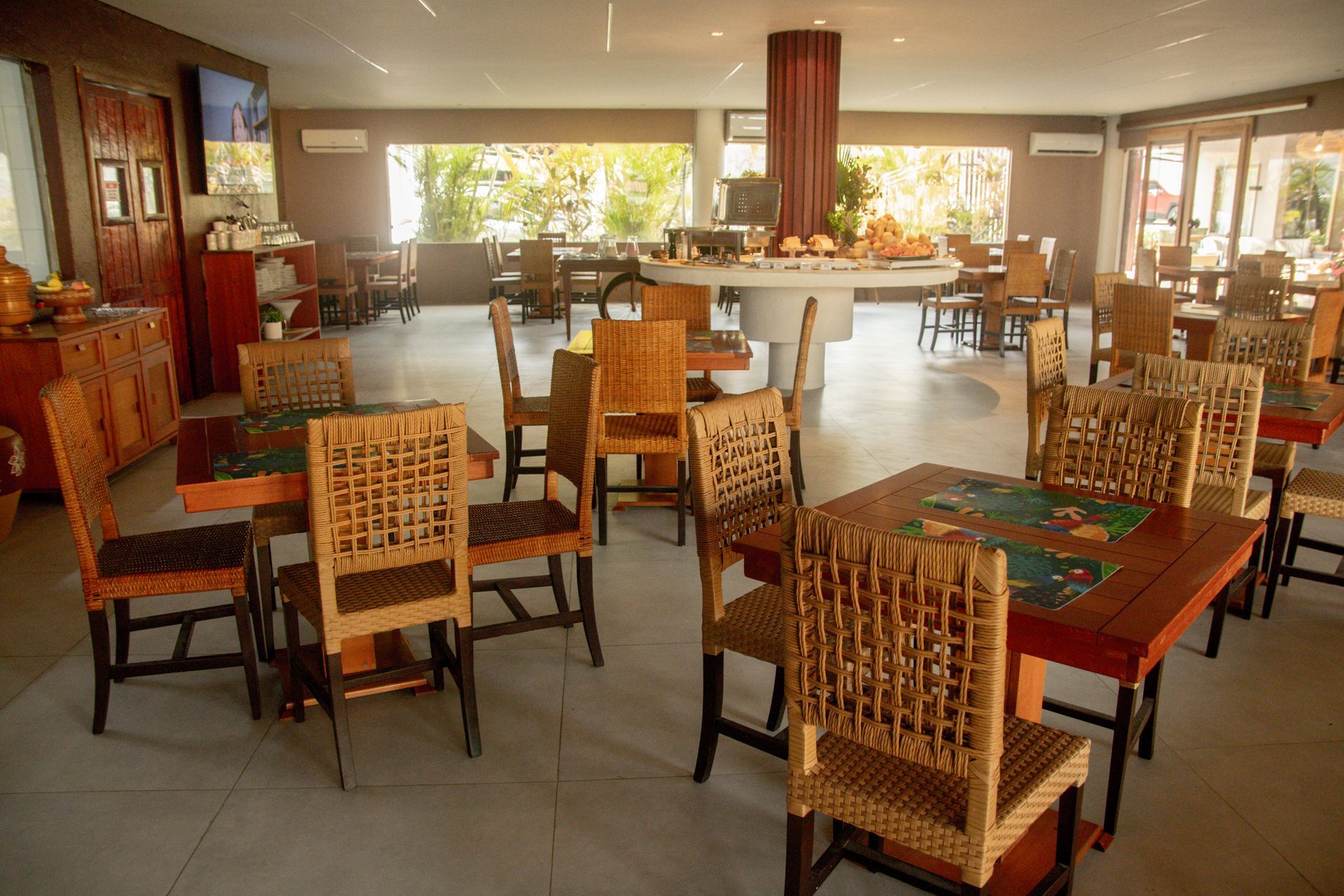Sala de jantar aconchegante com cadeiras de vime, mesas de madeira e grandes janelas com vista para a vegetação.