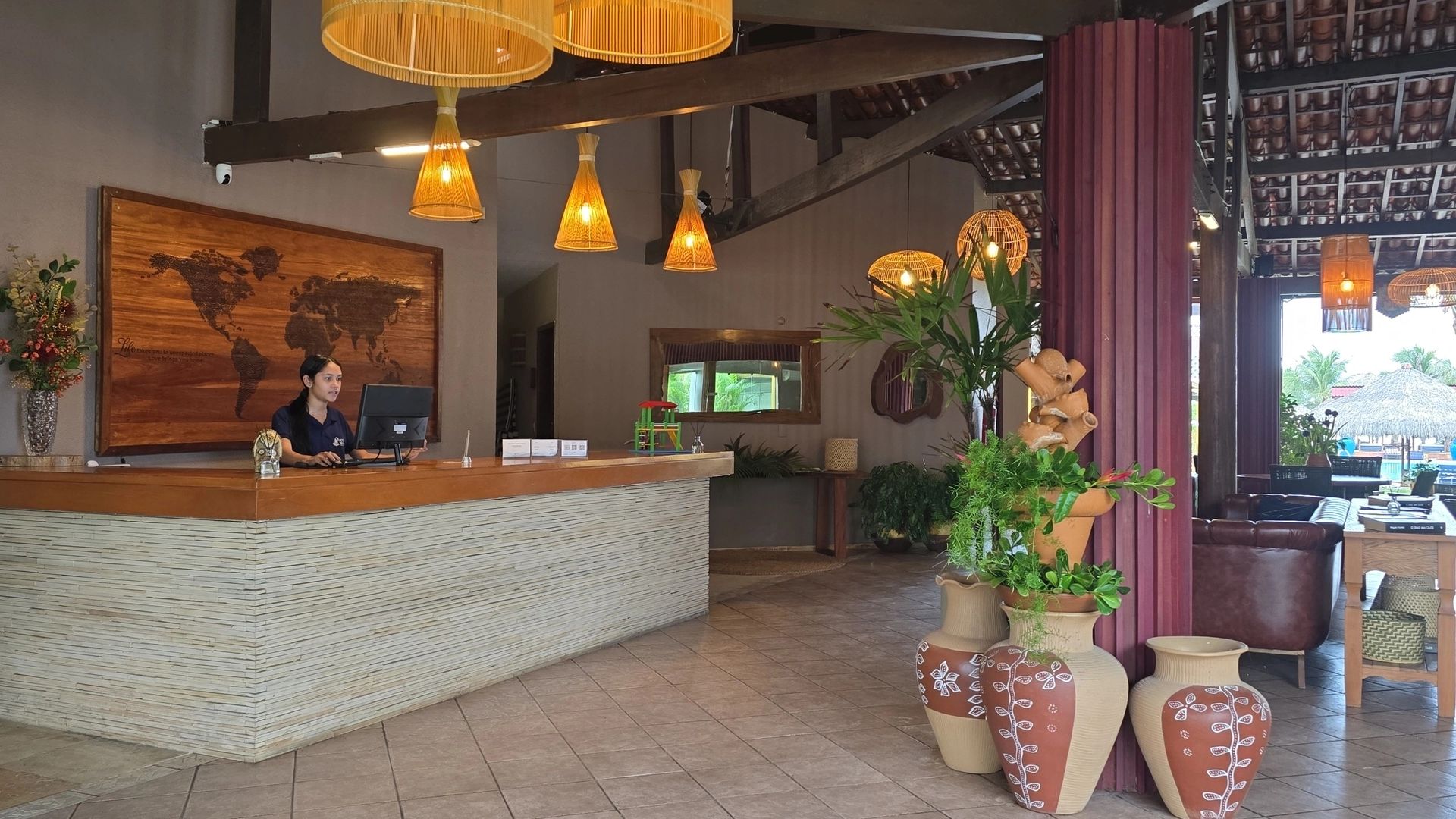 Hall de entrada do hotel com balcão de pedra branca, luminárias pendentes aconchegantes e plantas em vasos perto de uma área de estar.