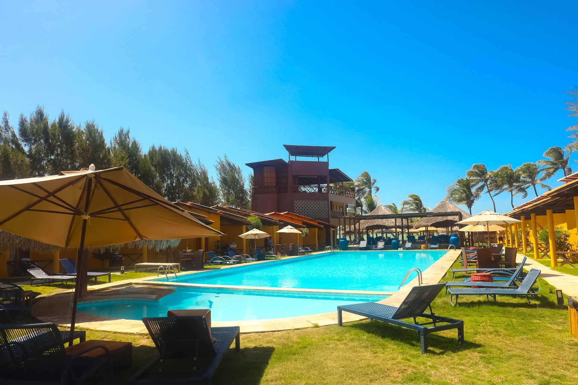 Piscina do resort com espreguiçadeiras, guarda-sóis e uma estrutura de madeira sob um céu azul brilhante.