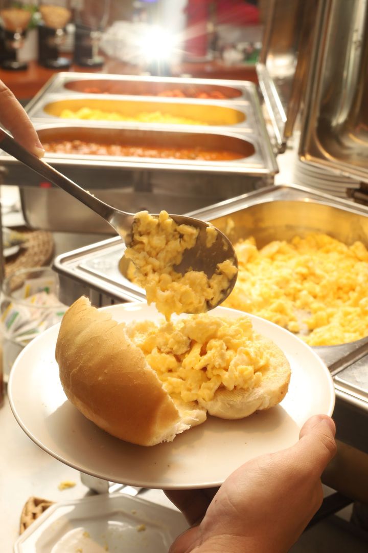 Uma mão segurando um prato com arroz e pão em um buffet com bandejas de comida.
