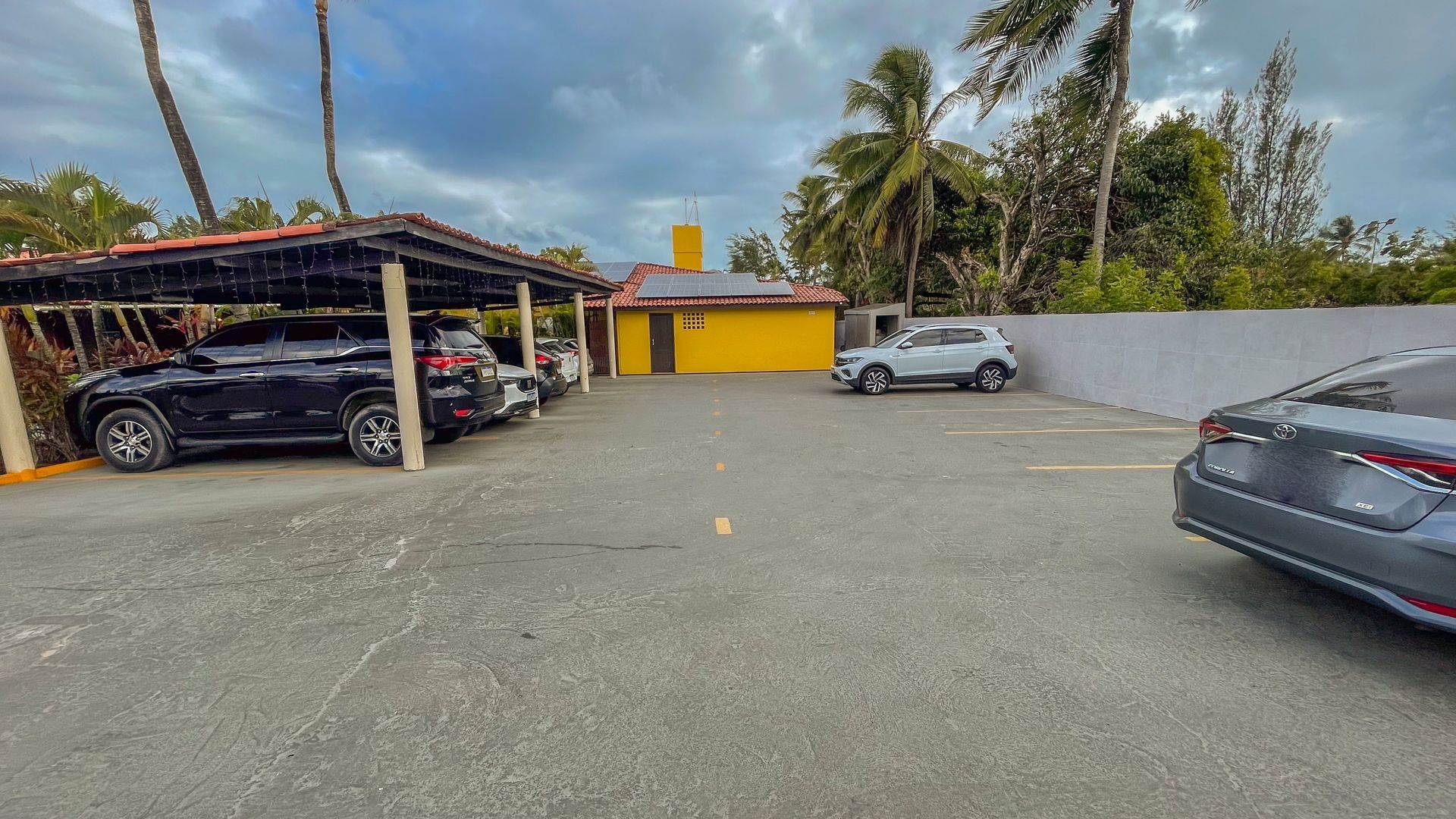 Estacionamento com vários carros, uma garagem coberta à esquerda e um prédio amarelo nos fundos.