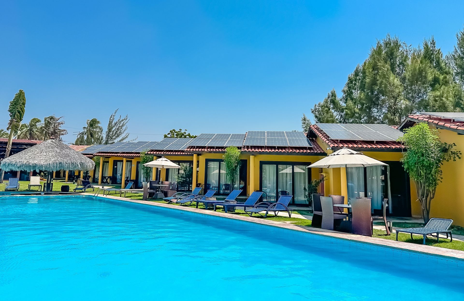 Piscina do resort com água azul, espreguiçadeiras e cabanas sombreadas sob um céu claro.