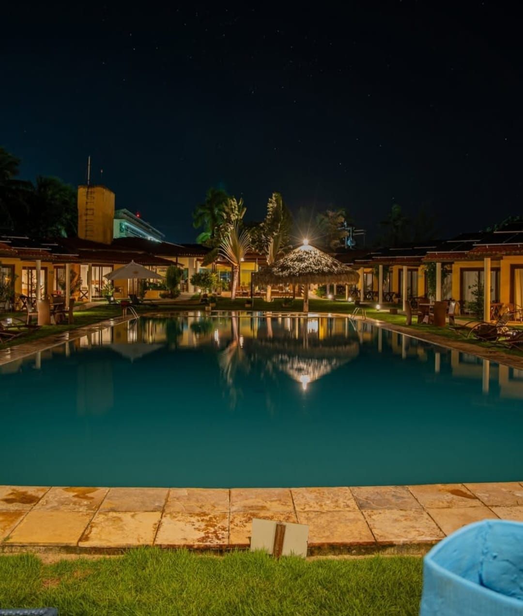 Piscina noturna do resort com reflexos tranquilos, palmeiras iluminadas e espreguiçadeiras ao longo do deck.