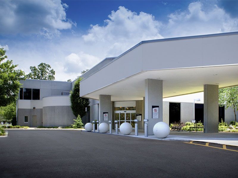 Exterior of a modern, light-colored building with a covered entrance, gray asphalt driveway, and blue sky.