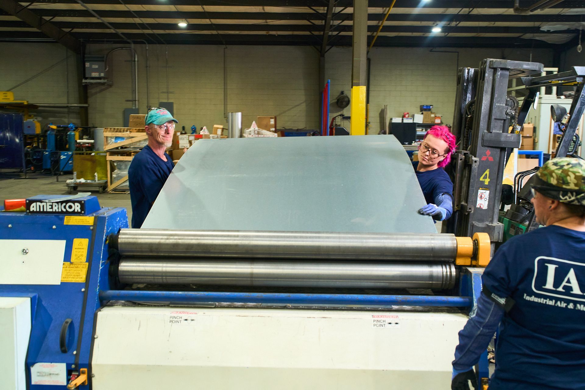 Three people rolling metal sheet using industrial machinery inside a factory.