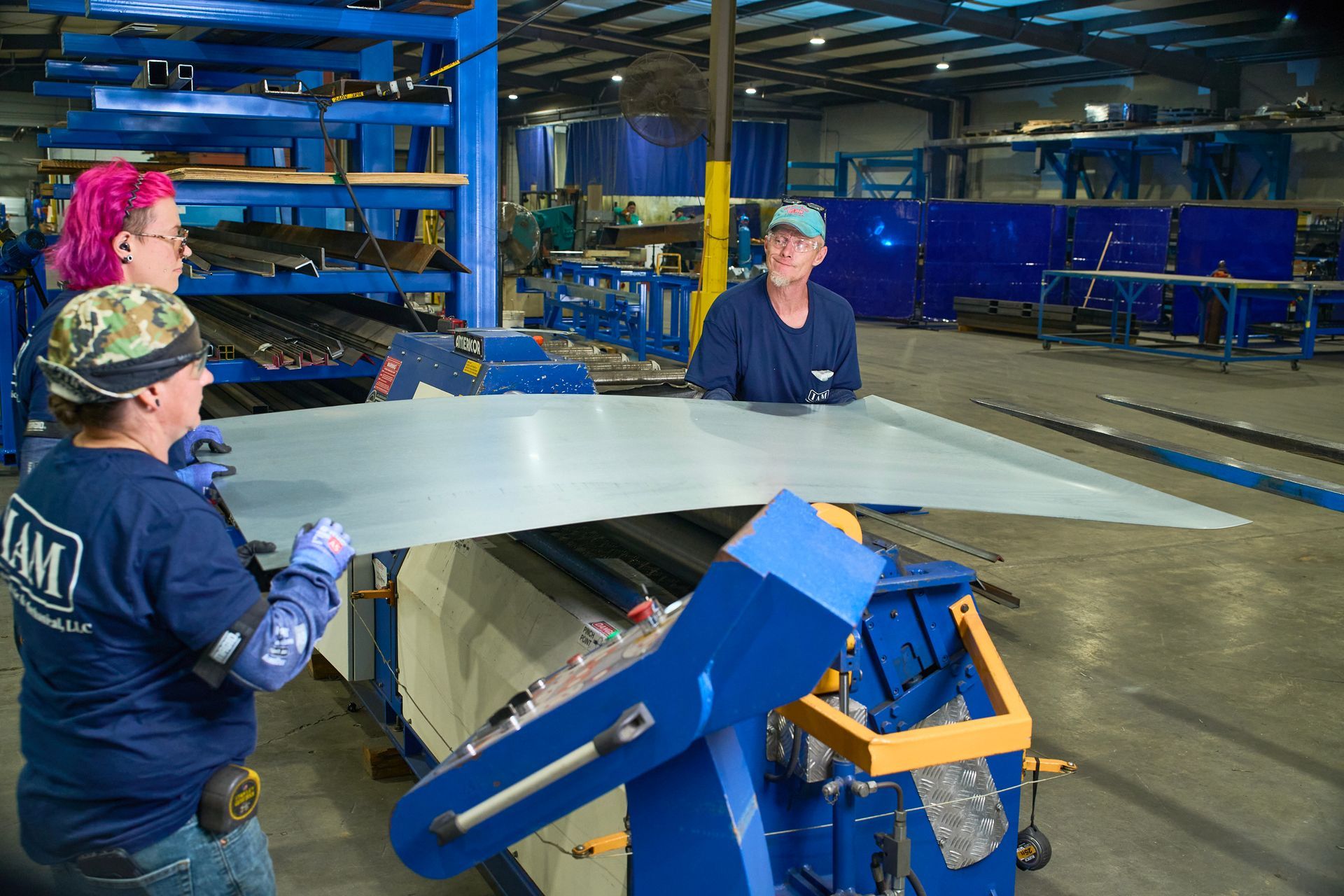 Three workers at a metal fabrication shop manipulating a large sheet of metal near a machine.
