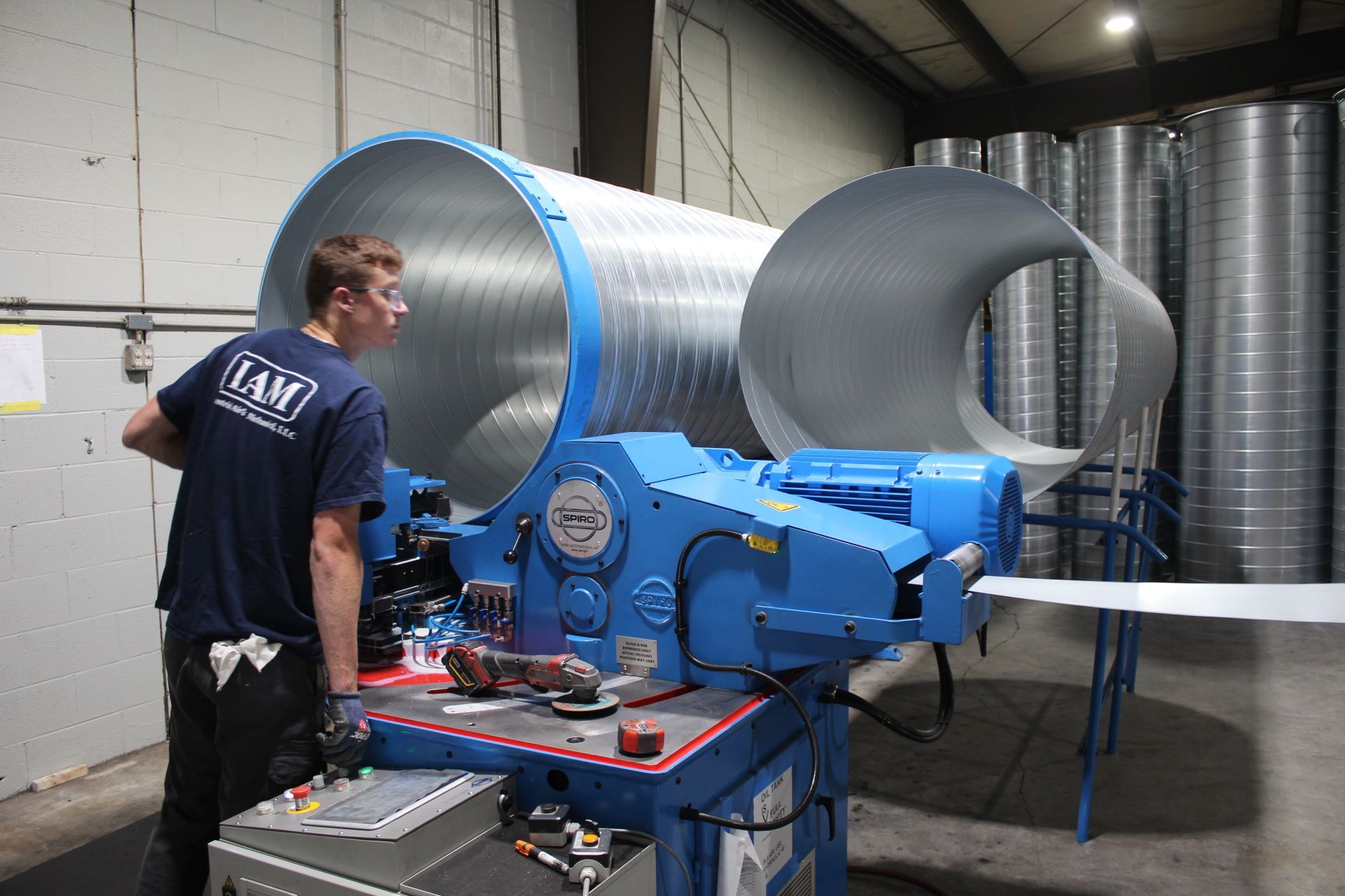 Man operating a blue machine to shape metal ductwork in a warehouse.