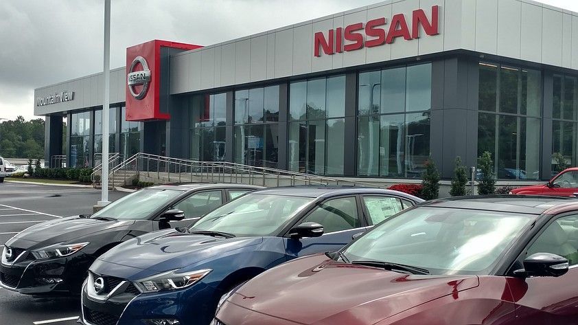 Nissan dealership exterior with parked cars in front. Red and silver building with large glass windows.