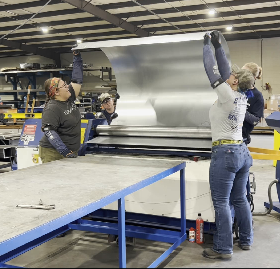 Workers in a metal shop feed a large sheet of metal into a rolling machine.