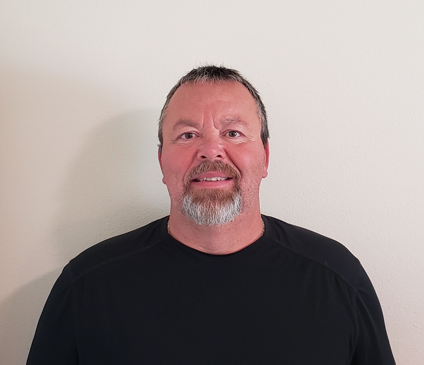 Man wearing a cap and dark blue shirt smiles at the camera, standing in front of a plain wall.