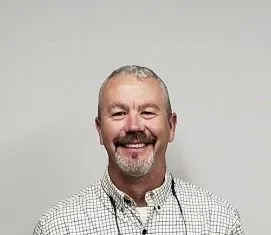 Man with a graying goatee smiles in a checkered shirt against a light gray background.