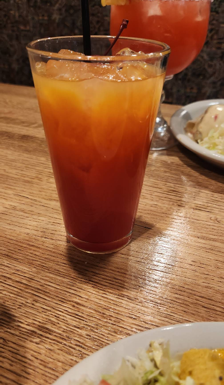 Orange-red layered cocktail with ice in a tall glass on a wooden table, other drinks and food visible.