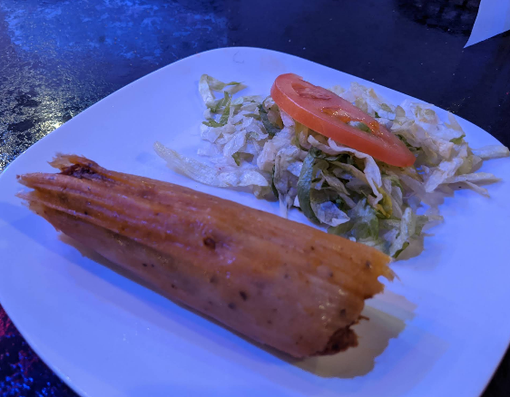 Tamale on a white plate, served with lettuce and a tomato slice.