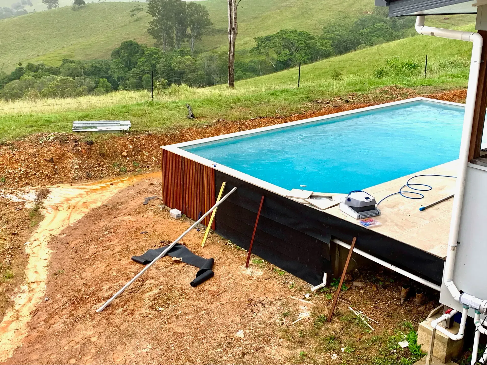 A partially built rectangular pool with blue water next to a building on a grassy hill. Construction equipment visible. — HinterCoast Pools In Curra, QLD