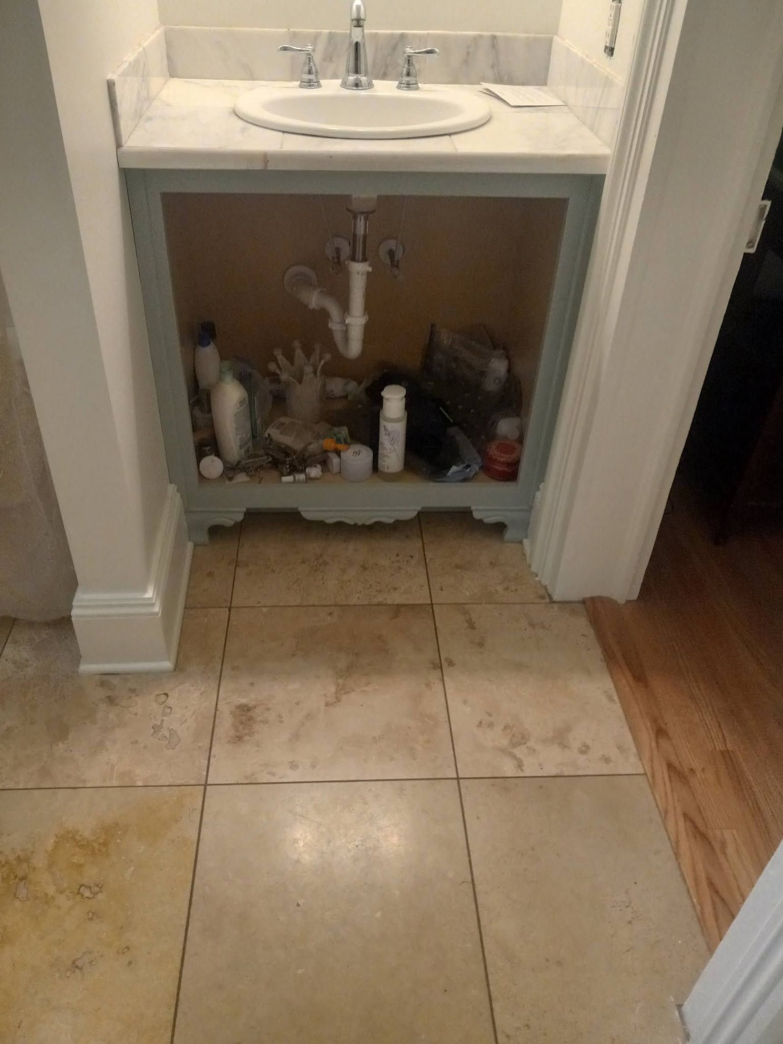 Bathroom sink cabinet with open storage, showing pipes and cleaning items underneath.