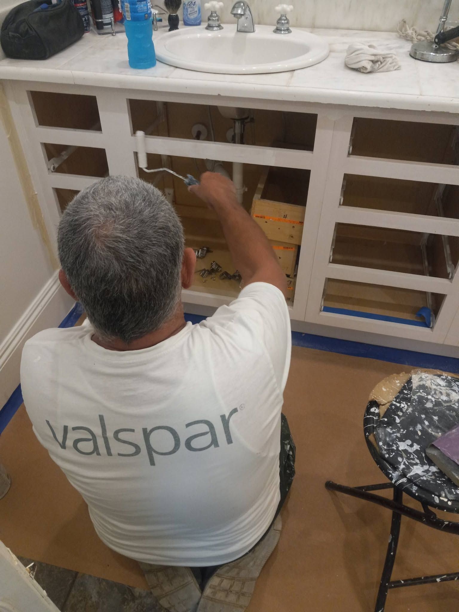 Person in a white Valspar shirt painting white bathroom cabinet doors under a sink.