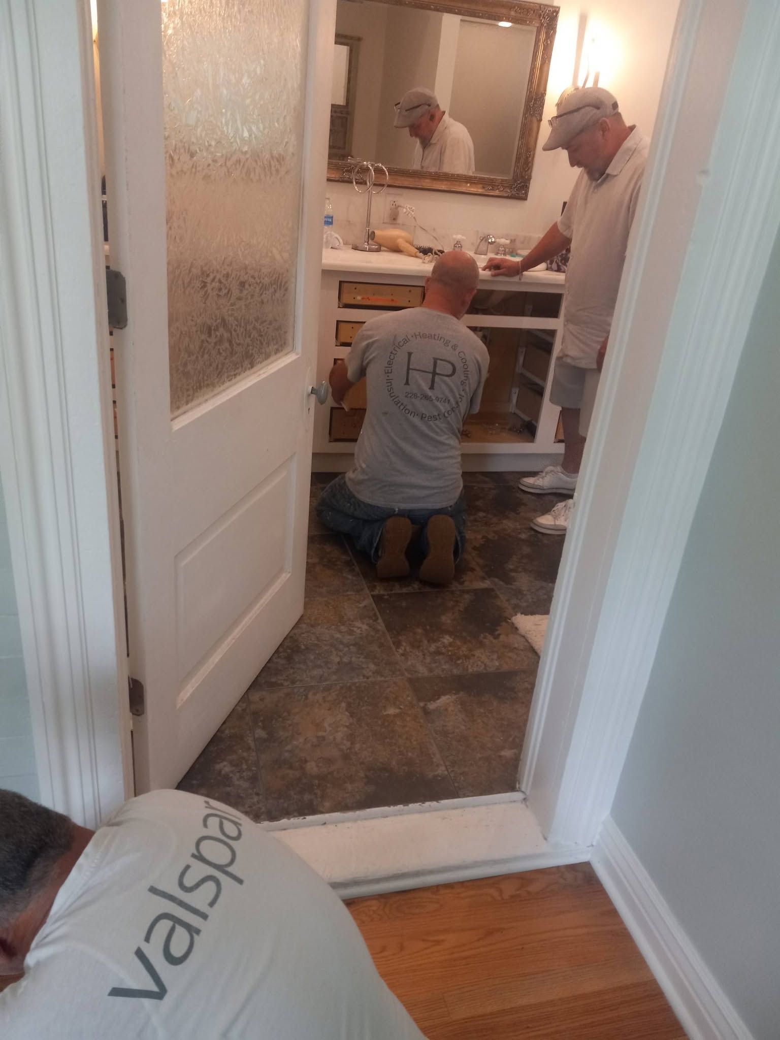 Two people kneel in a bathroom, working under a sink during a renovation.