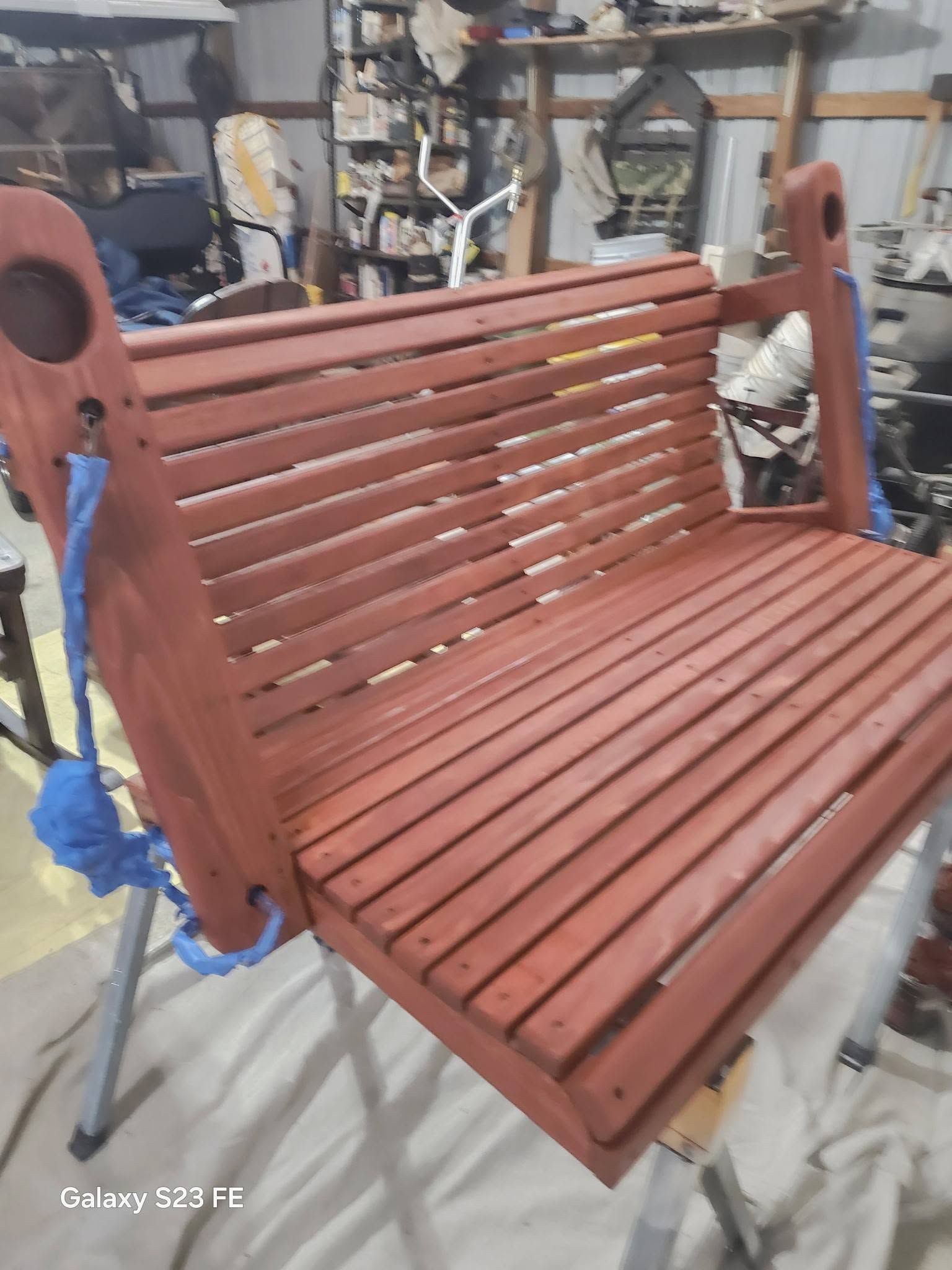 Red wooden bench swing seat hanging in a workshop, with unfinished surfaces and blue straps.