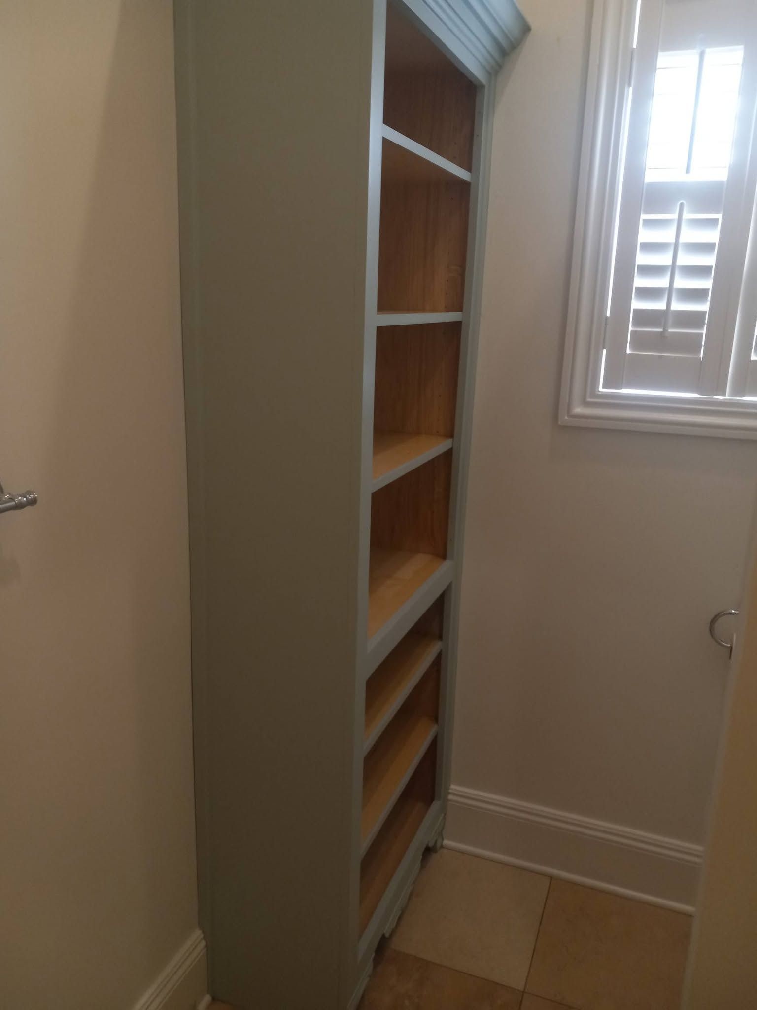 Tall built-in wooden shelf in a small tiled room beside a window and light-colored wall.