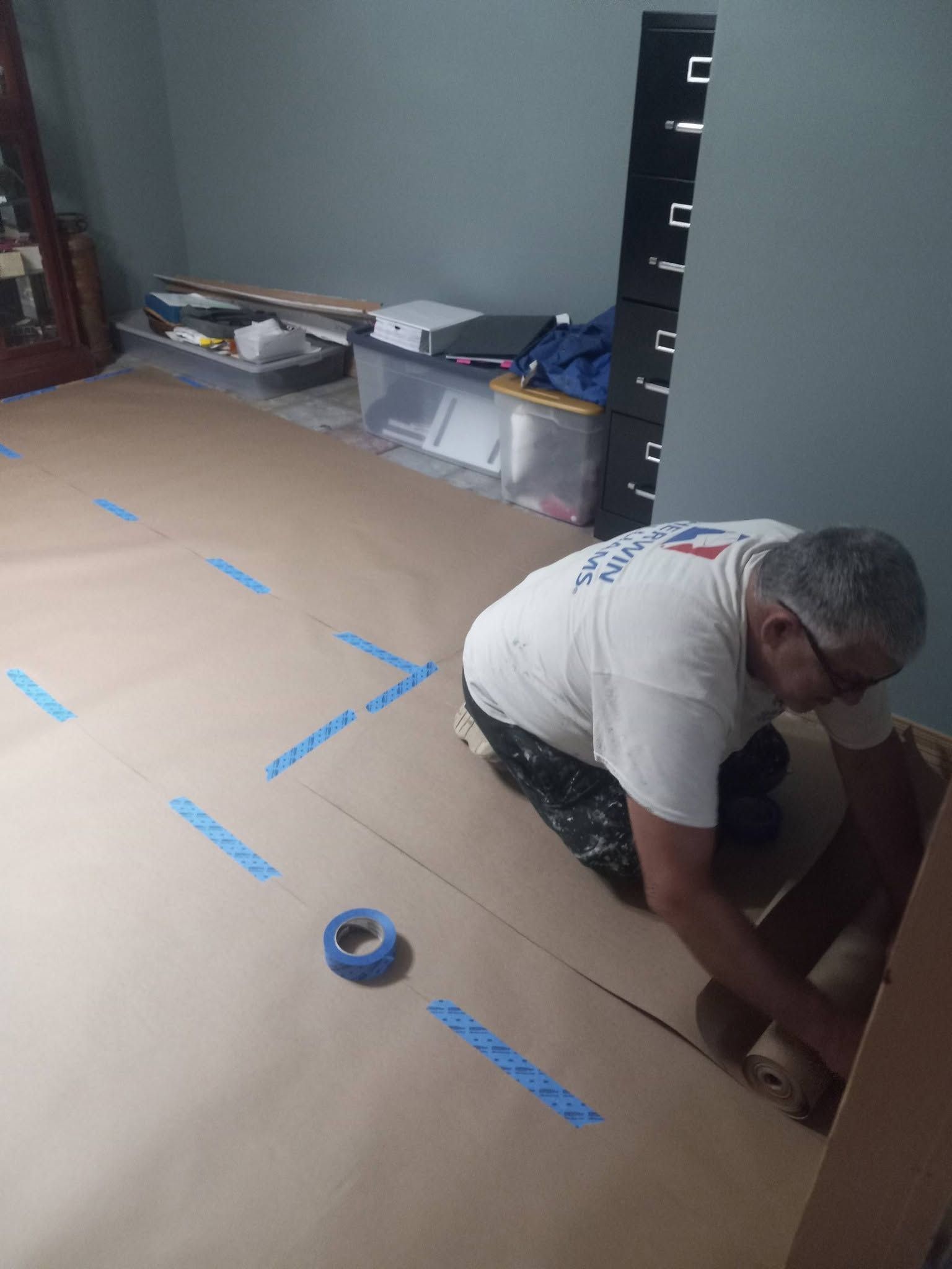 Person kneels on a taped floor, measuring or marking a large cardboard sheet in a room.