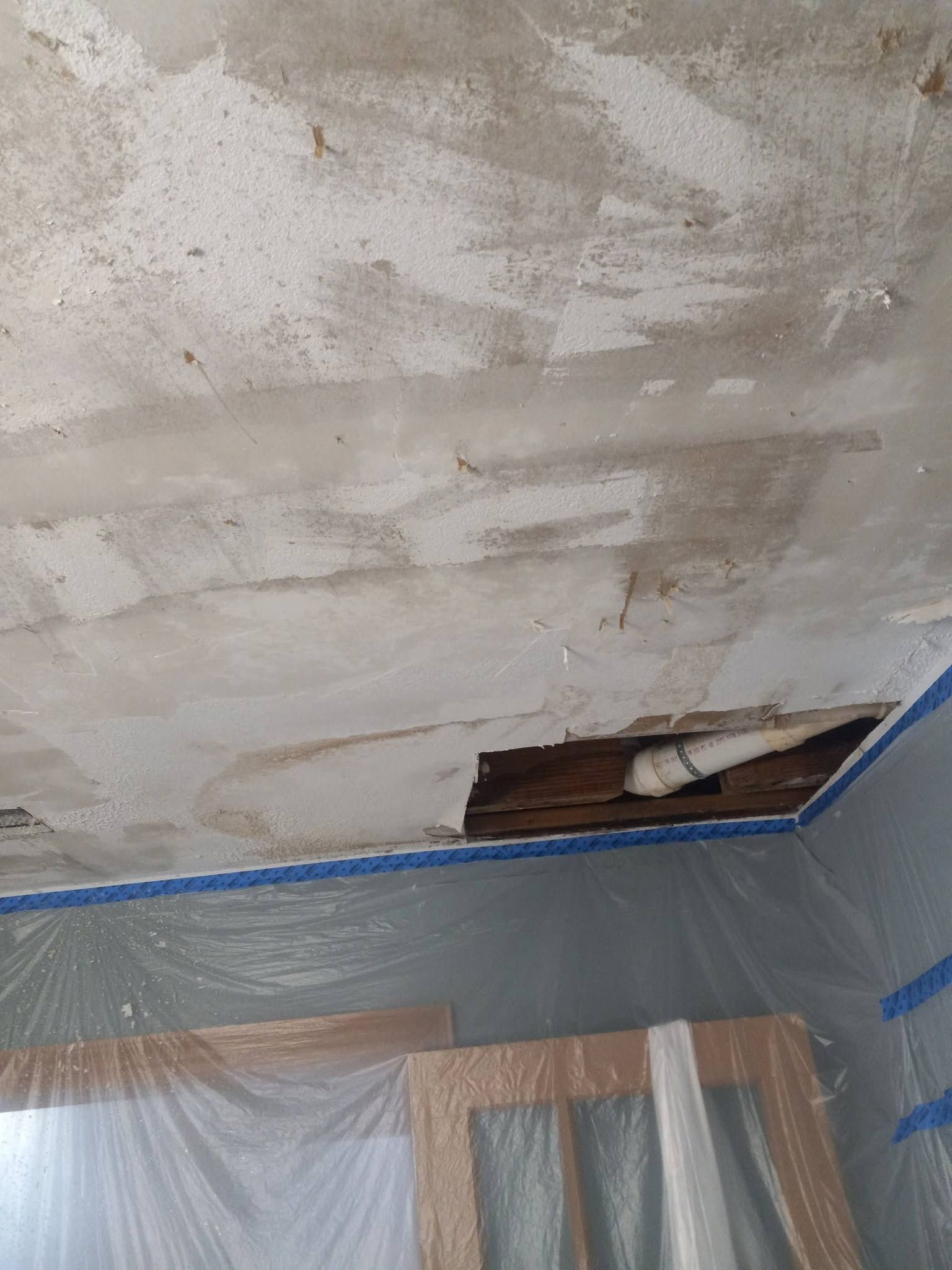 Damaged ceiling with peeling plaster and a hole, showing exposed wood above a room under plastic sheeting.