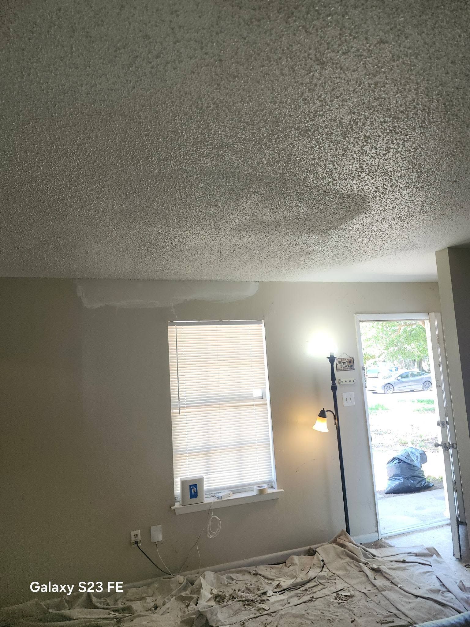 Ceiling under repair in a room with debris on the floor, open door, window, and lamp light nearby
