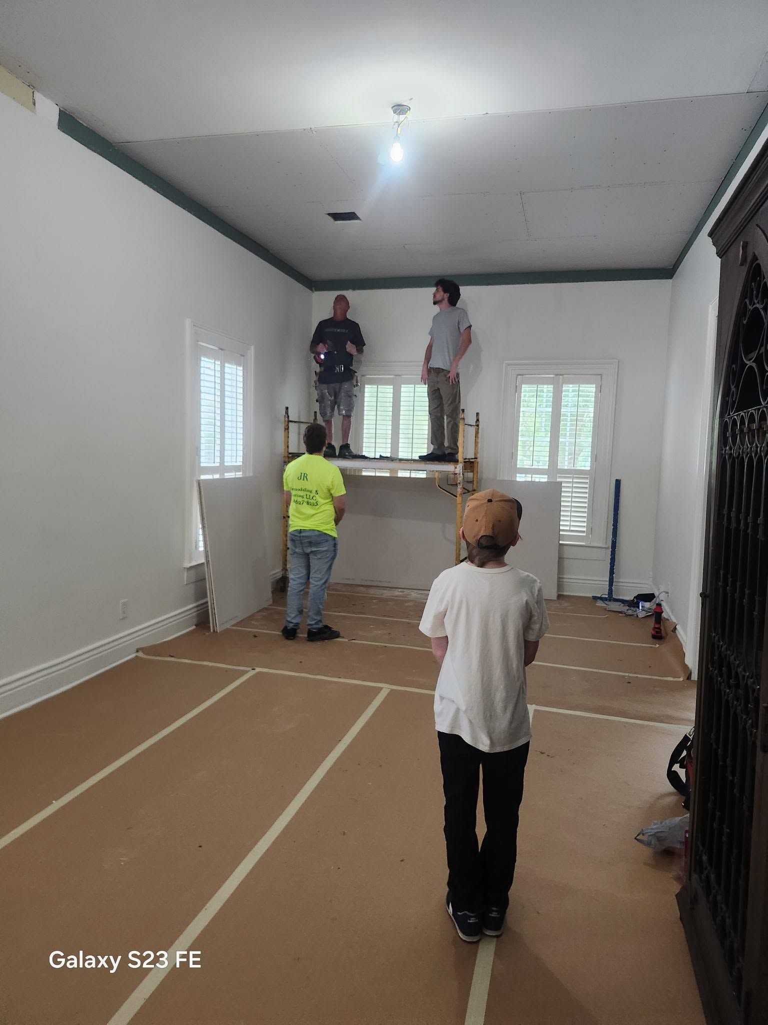 Workers installing a ceiling in an empty room with taped floor protection and ladders.