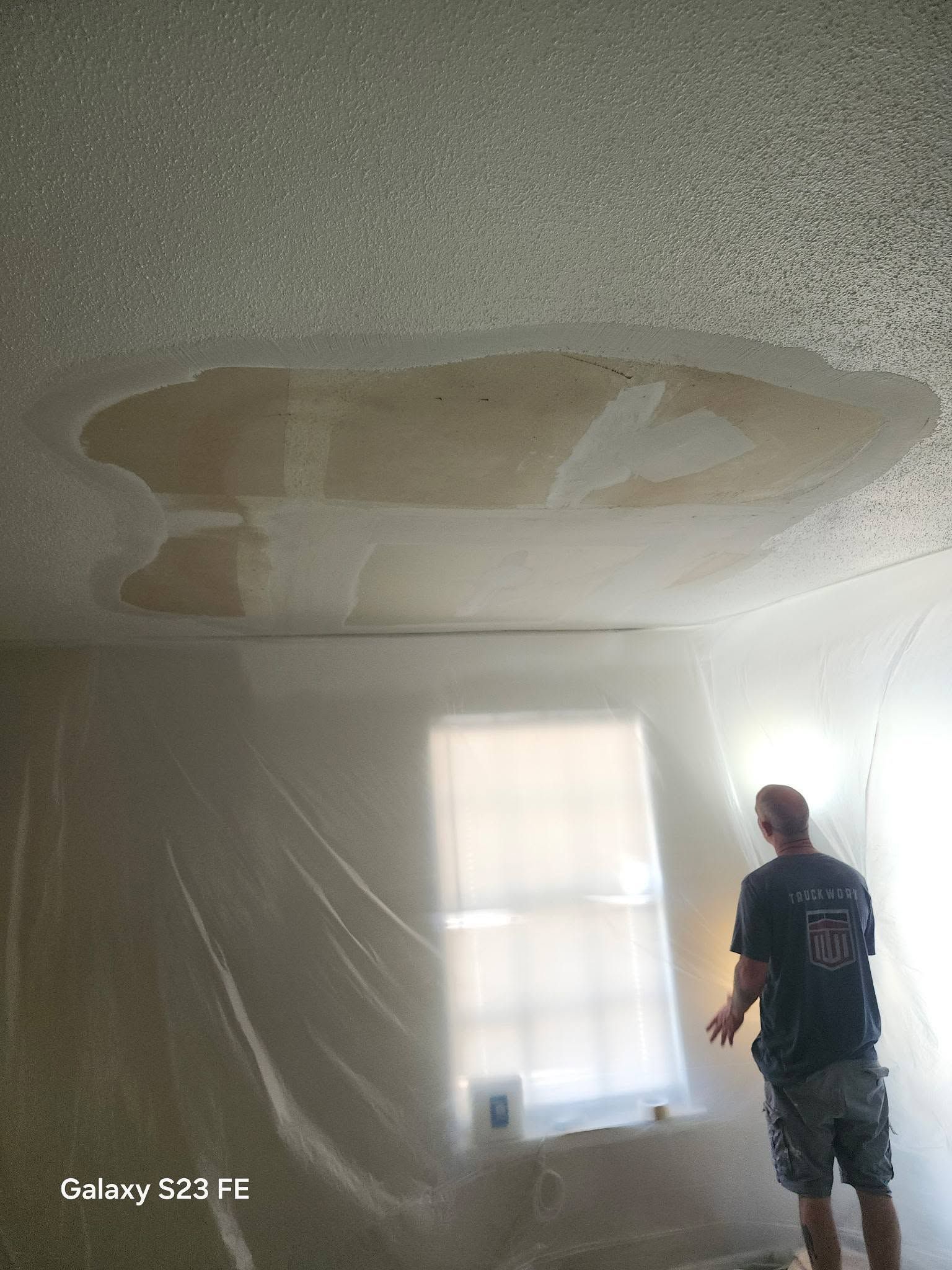 Person standing in a room with a large water stain on the ceiling near a window