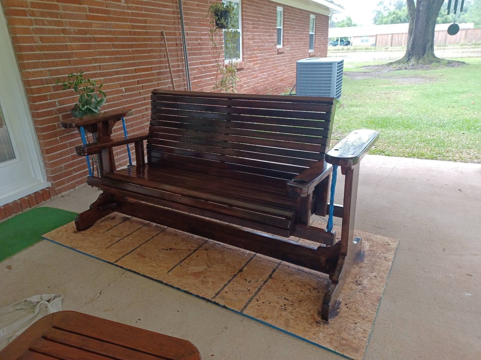 Wooden porch swing bench on a covered patio beside a brick wall and grassy yard