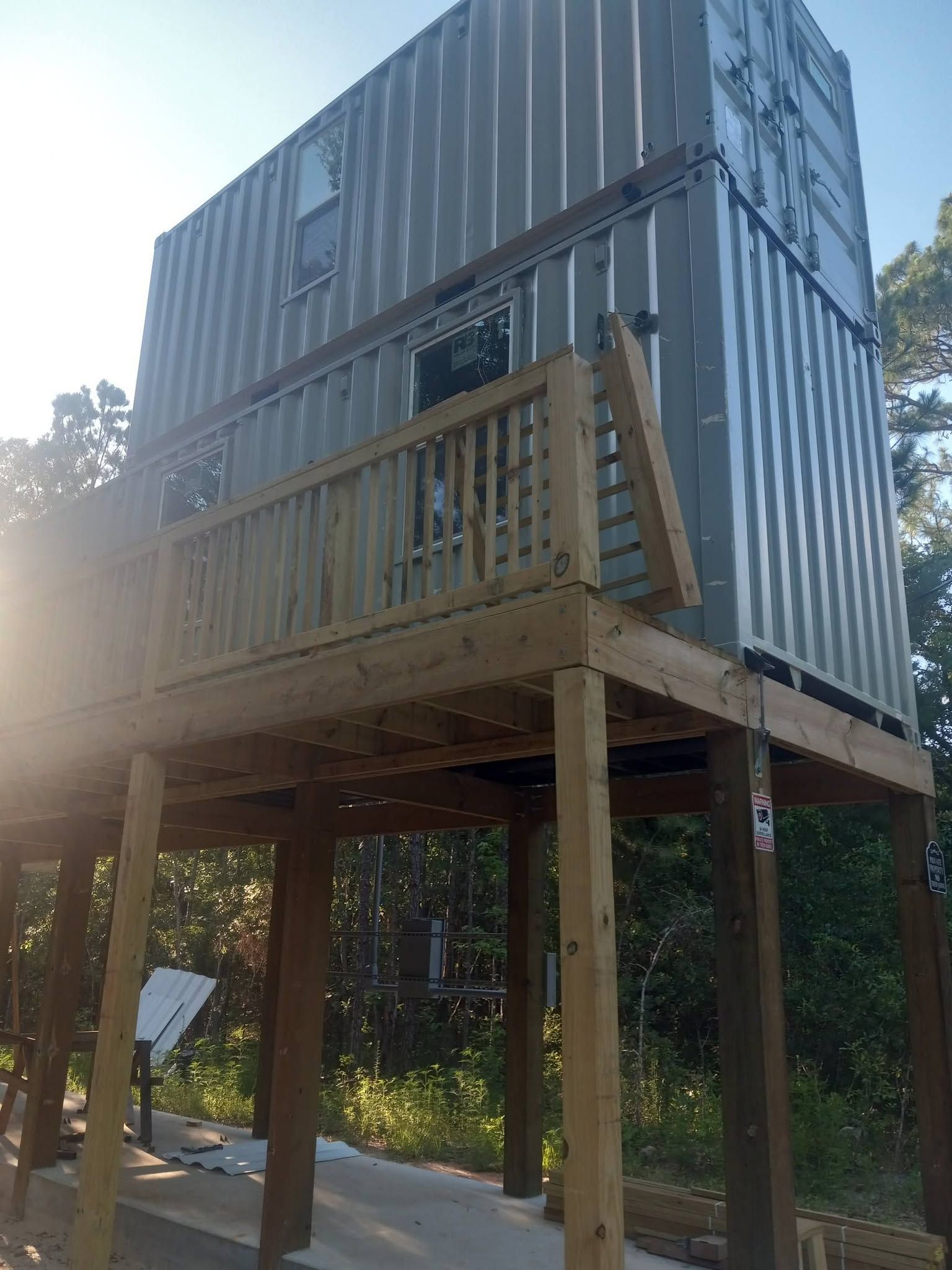 Raised container cabin on stilts with a wooden balcony in a wooded outdoor setting