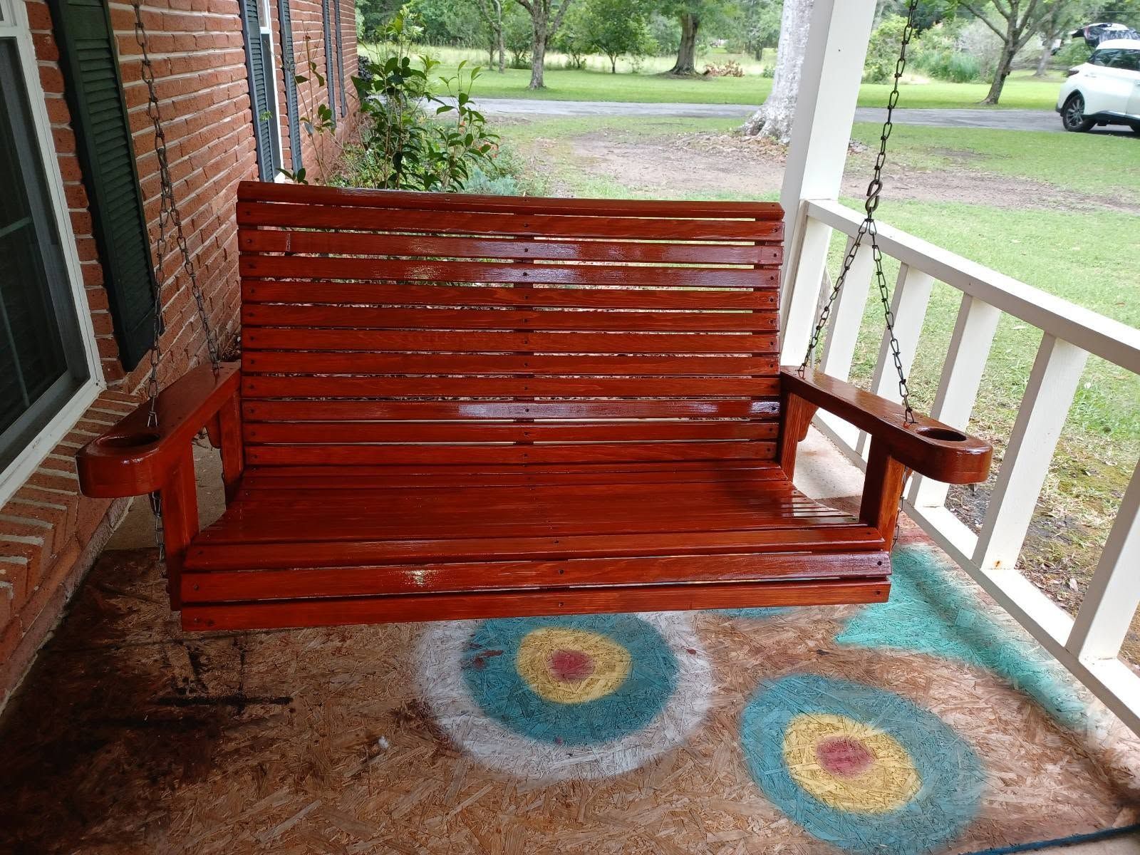 Red wooden porch swing hanging on a covered porch beside a white railing and colorful circular rug.