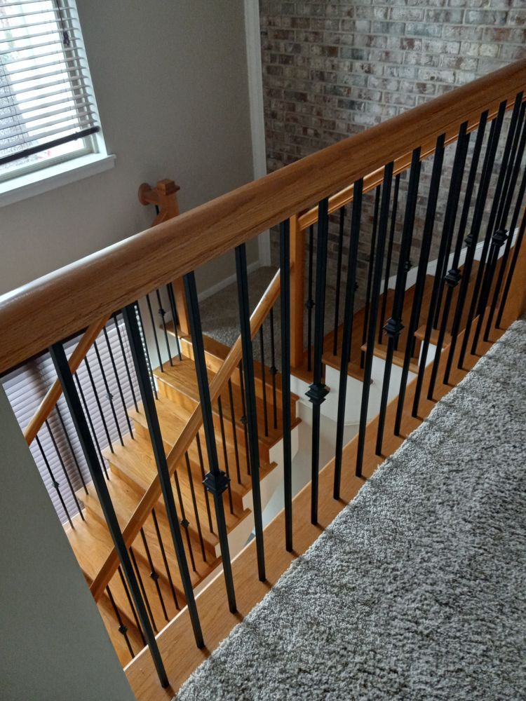 Staircase landing with wooden railing, black balusters, and carpeted stairs beside a brick wall and window.