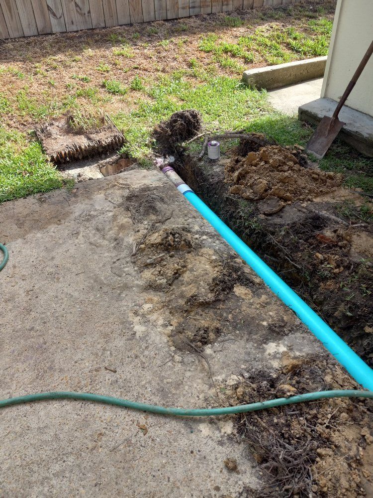 Backyard with a dirt trench beside a concrete patio, exposed blue pipe, and a green hose across the ground