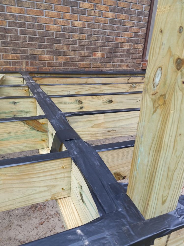 Wood deck framing with joists and black waterproof tape beneath a brick wall and wooden post.