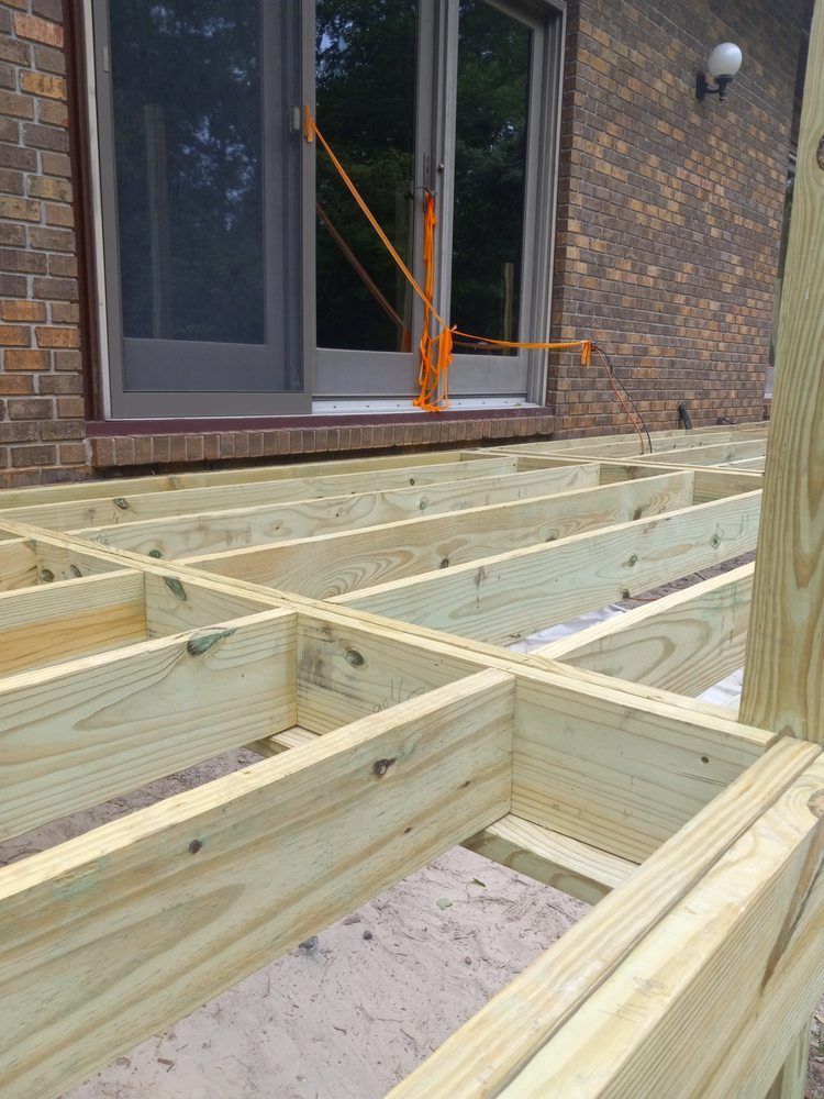 Wooden deck framing under construction beside a brick house and glass door, with orange straps nearby