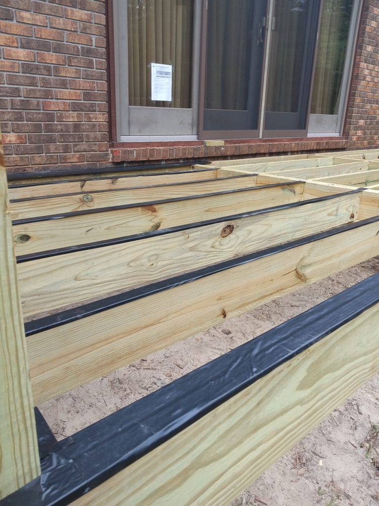 Partially built wooden deck beside a brick house, with black waterproofing strips between the boards