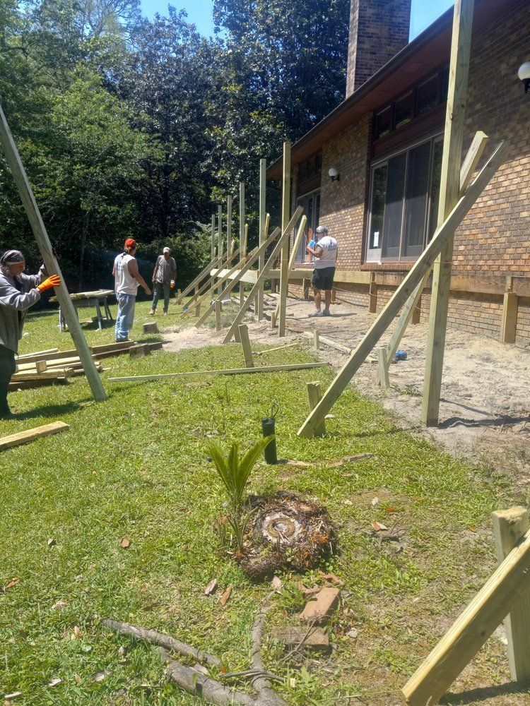 People working outdoors beside a house, setting wooden posts in a sunny yard.