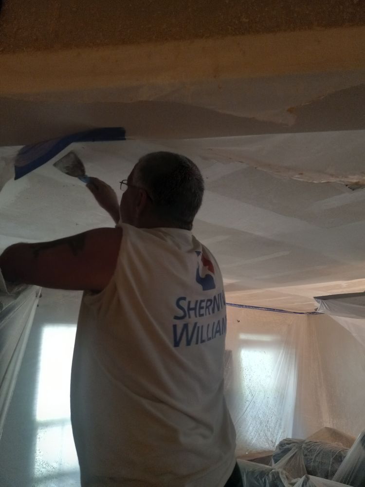Worker applying blue tape to a ceiling in a room with covered windows and furniture underneath