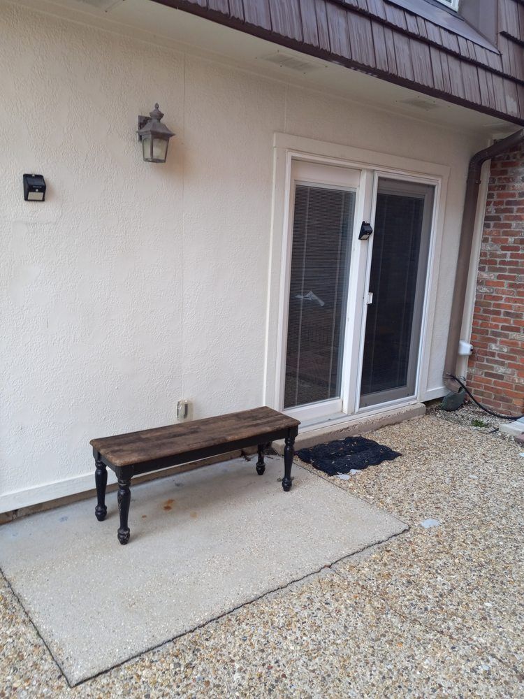 Covered patio with white stucco wall, sliding glass door, wall lantern, and a wooden bench on a concrete slab.