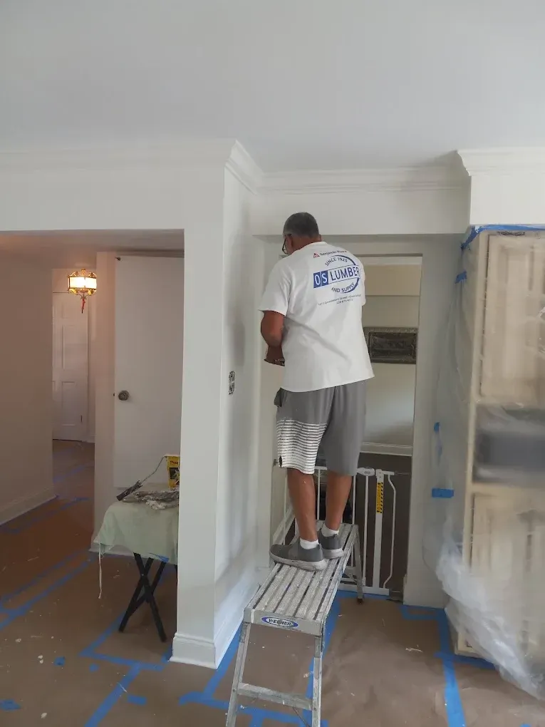 Worker standing on a ladder painting a white interior wall in a room under renovation