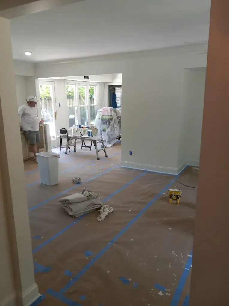 Home renovation scene with workers, taped-off floor, and painting supplies in a bright living room.