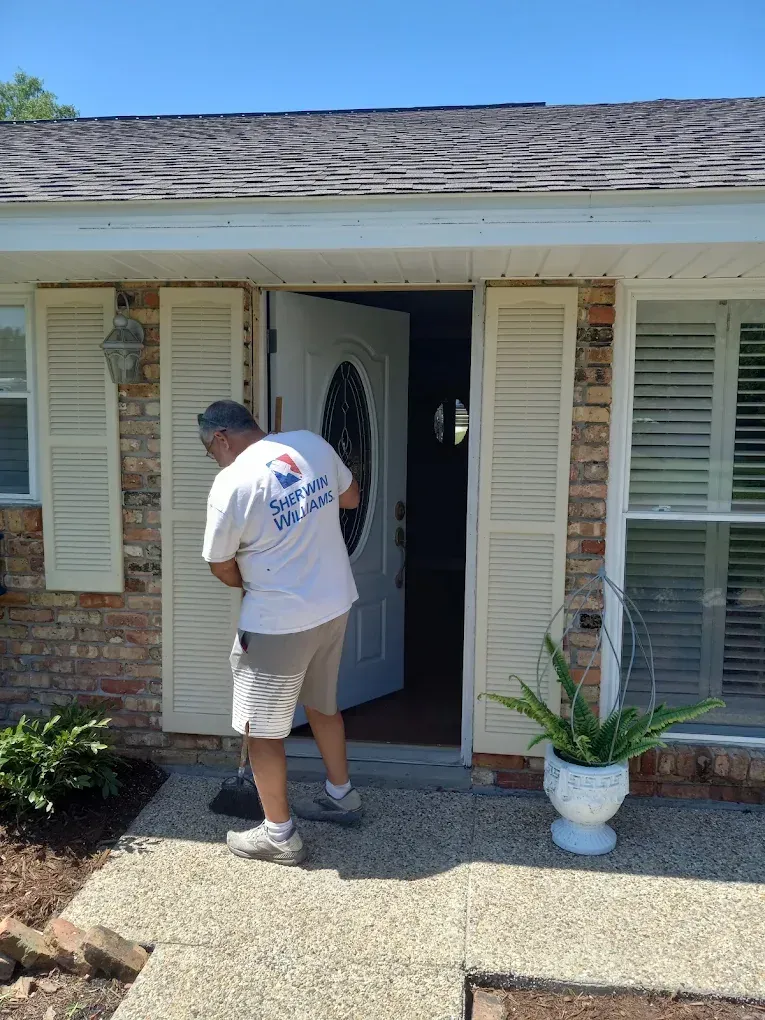 Man carrying a white door into a house entrance with open shutters and a potted plant on the porch