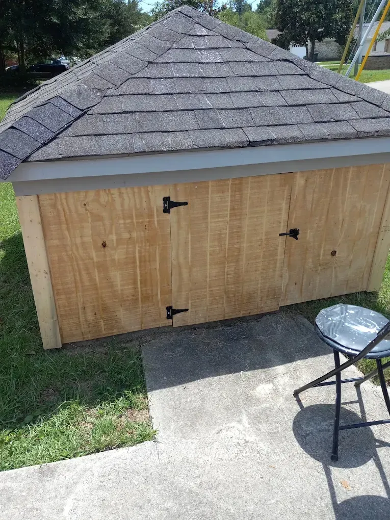 Wooden shed with a gray shingled roof beside a concrete walkway and grass, with a metal chair in the foreground