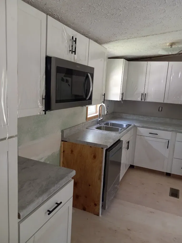 Bright kitchen with white cabinets, stainless sink, built-in microwave, and unfinished wooden base cabinet.