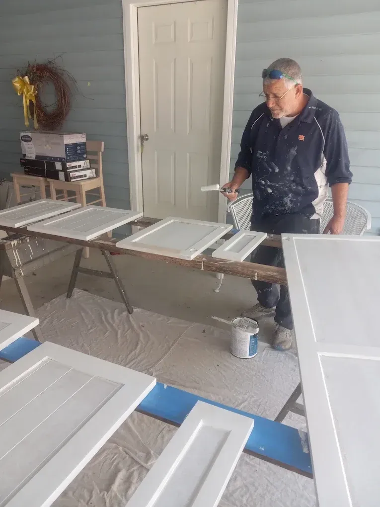 Person spray-painting white cabinet doors in a workshop with drop cloths and painting tools