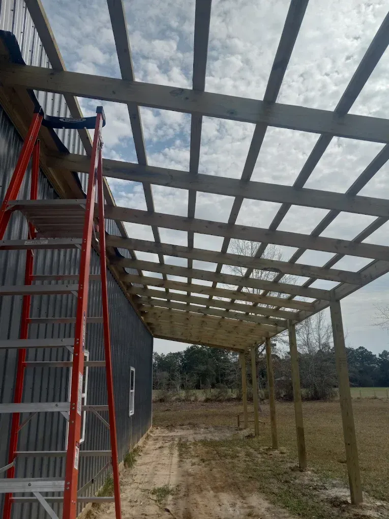 Under-construction covered walkway with red scaffolding beside a building and open field beyond