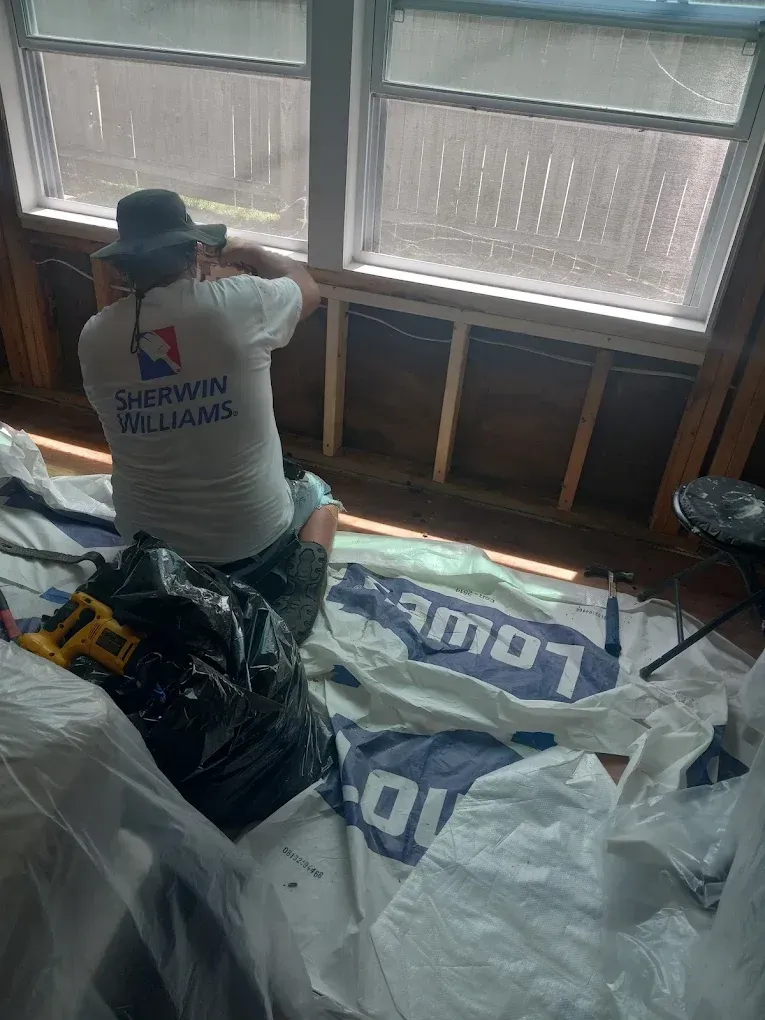 Worker installing insulation beneath windows in a room, with bags of insulation materials on the floor.