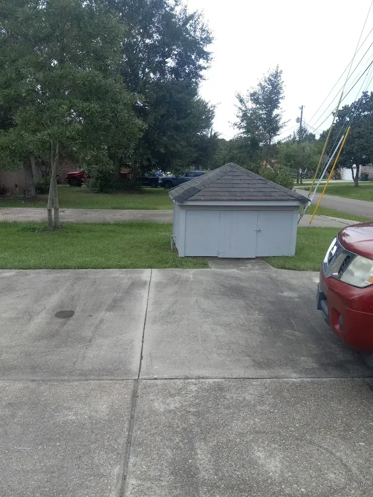 Driveway beside a small gray shed, with a red car parked at the right and trees in the yard.